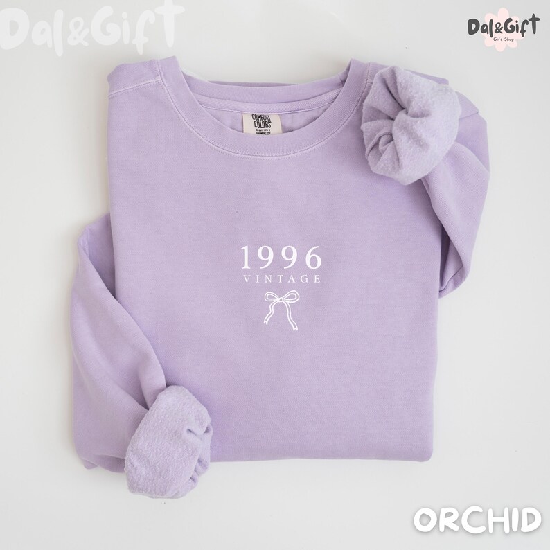 May include: A light purple sweatshirt with the text "1996 VINTAGE" and a bow graphic. The sweatshirt is a solid color and has a crew neck. The sleeves are cuffed. The word "ORCHID" is at the bottom right.