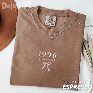 May include: A brown short-sleeve t-shirt with the text "1996 VINTAGE" and a bow graphic. The shirt has a crew neck and a silver necklace with a pendant. The words "SHORT SLEEVE ESPRESSO" are at the bottom.