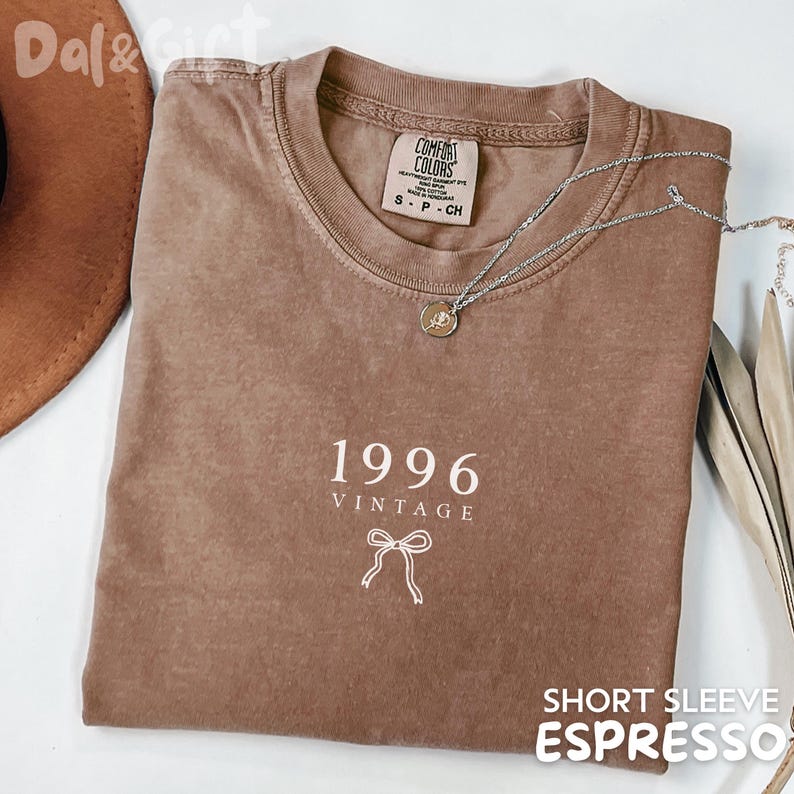 May include: A brown short-sleeve t-shirt with the text "1996 VINTAGE" and a bow graphic. The shirt has a crew neck and a silver necklace with a pendant. The words "SHORT SLEEVE ESPRESSO" are at the bottom.