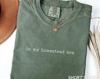 May include: A moss green short-sleeve t-shirt with the phrase "in my homestead era" printed in white. A gold necklace with a pendant is draped over the shirt. The shirt has a Comfort Colors tag.