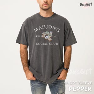 May include: Dark gray short-sleeve t-shirt with the text "MAHJONG SOCIAL CLUB EST 1923" in white. The shirt features a graphic of Mahjong tiles. The shirt is a pepper color.