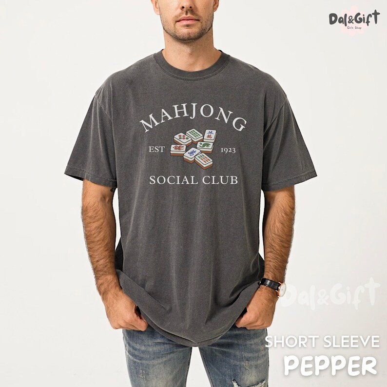 May include: Dark gray short-sleeve t-shirt with the text "MAHJONG SOCIAL CLUB EST 1923" in white. The shirt features a graphic of Mahjong tiles. The shirt is a pepper color.