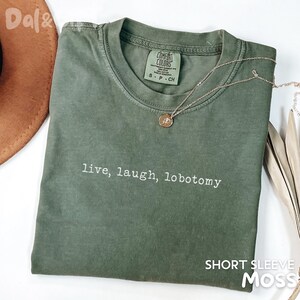 May include: A moss green short-sleeve t-shirt with the phrase "live, laugh, lobotomy" printed in white. The shirt has a crew neck and a Comfort Colors tag. A gold necklace with a pendant is draped on the shirt.