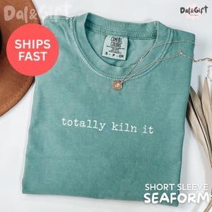 May include: Seafoam green short sleeve t-shirt with the phrase "totally kiln it" in white. A gold-colored necklace with a pendant rests on the shirt. The shirt has a Comfort Colors tag.