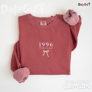 May include: Crimson-colored sweatshirt with the text "1996 VINTAGE" and a bow graphic. The sweatshirt has a crew neck and a soft, fuzzy lining at the cuffs. The sweatshirt is a solid color and is laid flat on a white surface.