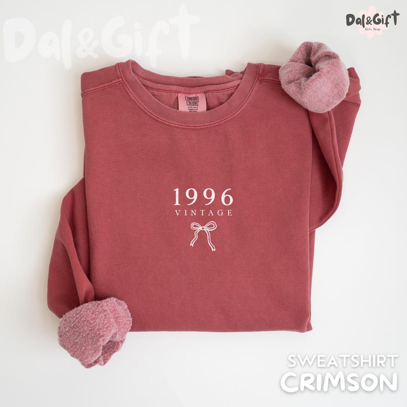 May include: Crimson-colored sweatshirt with the text "1996 VINTAGE" and a bow graphic. The sweatshirt has a crew neck and a soft, fuzzy lining at the cuffs. The sweatshirt is a solid color and is laid flat on a white surface.