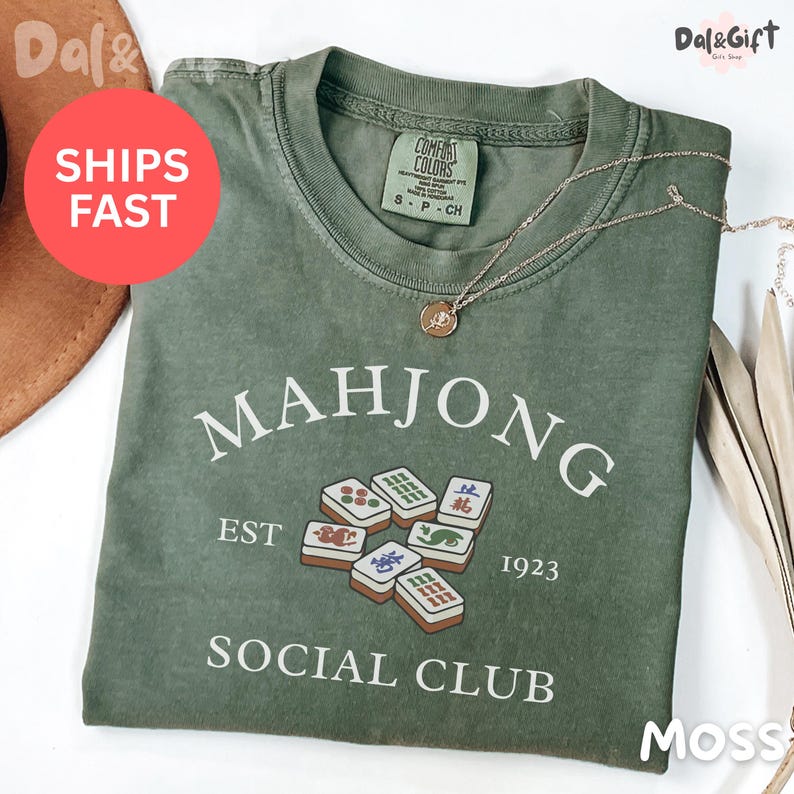 May include: Moss green t-shirt with the text "MAHJONG SOCIAL CLUB EST 1923" and a graphic of Mahjong tiles. A gold necklace with a pendant is draped across the shirt. The shirt is folded and has a tag that says "COMFORT COLORS".