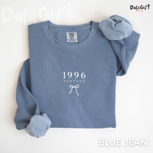 May include: A blue jean-colored sweatshirt with the year "1996" and the word "VINTAGE" printed in white. A white bow graphic is below the text. The sweatshirt has a crew neck and long sleeves. A matching scrunchie rests on the shoulder.
