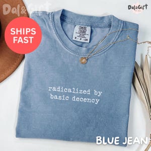 May include: A blue jean colored t-shirt with the phrase "radicalized by basic decency" in white text. The shirt has a crew neck and short sleeves. A gold necklace with a pendant rests on the shirt.