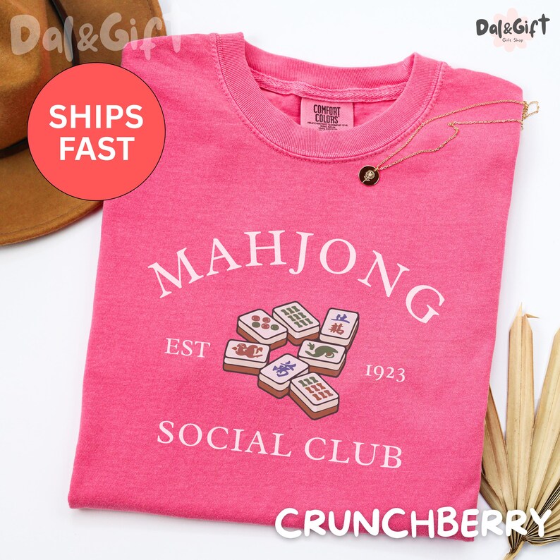 May include: A pink t-shirt with the text "MAHJONG SOCIAL CLUB EST 1923" and a graphic of Mahjong tiles. The shirt is a Comfort Colors brand. A gold necklace with a pendant is on the shirt.
