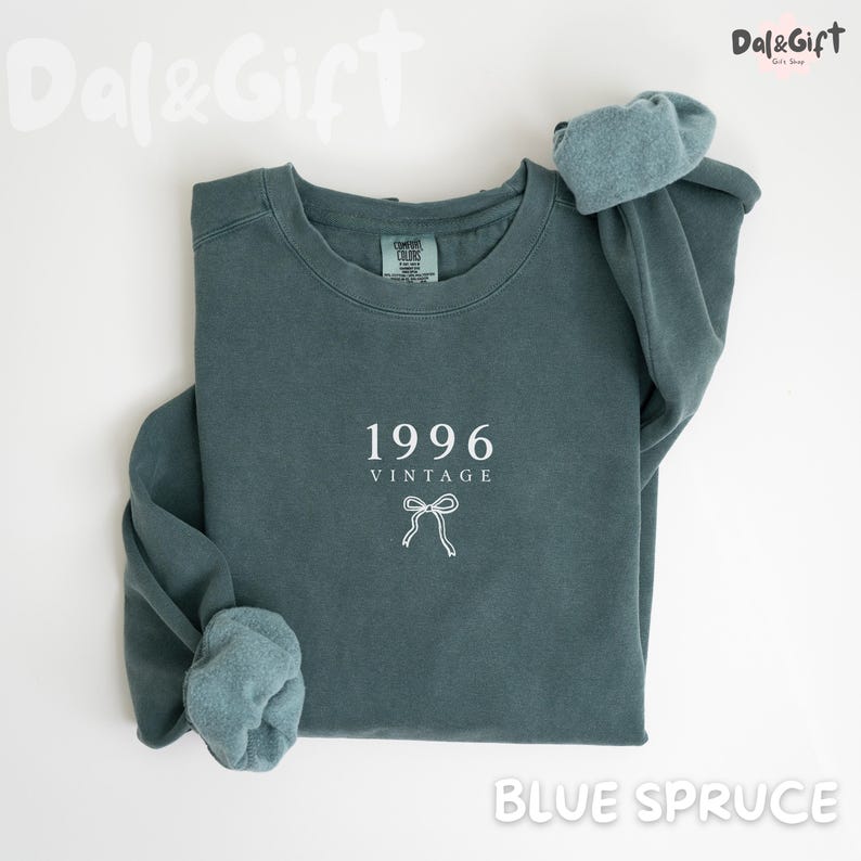 May include: A blue spruce sweatshirt with the year "1996" and the word "VINTAGE" printed in white. A white bow graphic is below the text. The sleeves are cuffed, and the sweatshirt is folded. The words "Dal&Gift" and "Blue Spruce" are also visible.