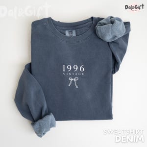 May include: A denim blue sweatshirt with the year "1996" and the word "VINTAGE" printed in white. A white bow graphic is below the text. The sleeves are cuffed, and the sweatshirt is lying flat on a white surface.