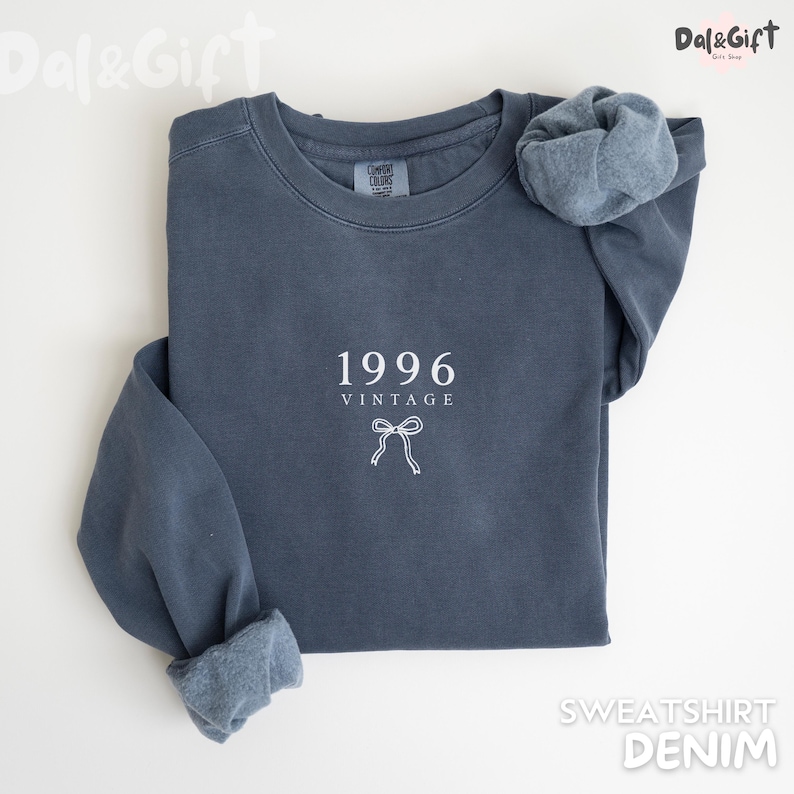 May include: A denim blue sweatshirt with the year "1996" and the word "VINTAGE" printed in white. A white bow graphic is below the text. The sleeves are cuffed, and the sweatshirt is lying flat on a white surface.
