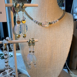 May include: A jewellery display featuring a necklace, bracelet, and earrings. The necklace and bracelet have a gold chain with clear and iridescent beads. The earrings are gold hoops with dangling iridescent and gold accents. The jewellery is displayed on a mannequin.