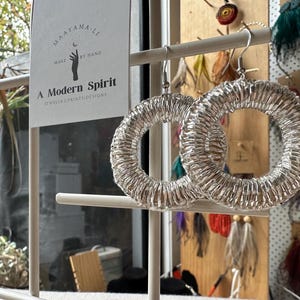May include: A pair of silver-toned hoop earrings with a textured, coiled wire design. The earrings are displayed on a white stand next to a white card featuring the text Maayama-Li, Make by Hand, A Modern Spirit, and Jewelry Prints Designs.