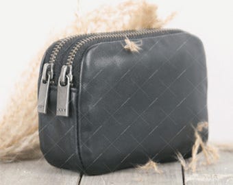 May include: A black leather cosmetic bag with two zippers and silver-toned pulls. The bag is rectangular and sits on a wooden surface. Dried ornamental grass is in the background.