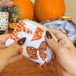 May include: A small, white, zippered pouch featuring a repeating pattern of orange and white checkered bows, brown and white polka-dot bows, and orange floral motifs. The pouch has a light beige zipper closure along the top edge.