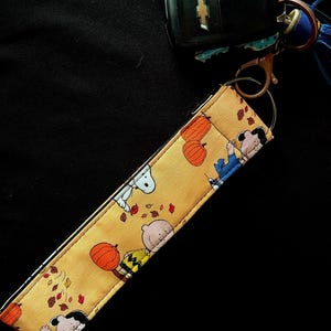 May include: A fabric wristlet keychain featuring a yellow background with a repeating pattern of Peanuts characters, orange pumpkins, and scattered autumn leaves. The wristlet includes a bronze-colored metal lobster clasp and a split ring for keys.
