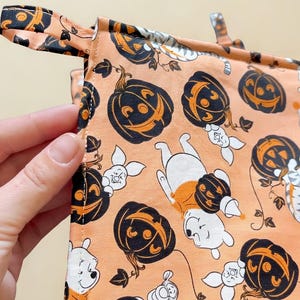May include: A square fabric pot holder featuring a light orange background with a repeating pattern of black jack-o'-lanterns and cartoon characters including a bear, a small pig, and a striped tiger. The item includes a fabric loop for hanging.