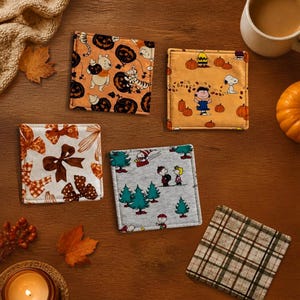 May include: Five square fabric coasters featuring various patterns: one with Winnie the Pooh and pumpkins, one with Peanuts characters in a pumpkin patch, one with brown bows, one with a Peanuts winter scene, and one with a green and brown plaid pattern.
