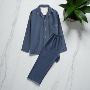 Può includere: Un completo pigiama a due pezzi blu denim. La parte superiore ha una chiusura con bottoni, una tasca sul petto e finiture bianche sul colletto e sui polsini. I pantaloni abbinati hanno una vita elasticizzata. Il set è appoggiato su una superficie di marmo bianco.