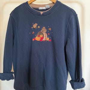 May include: A navy blue long-sleeve sweatshirt featuring a crew neckline with a tan inner collar. The center chest displays an embroidered design of a squirrel, a bird, pumpkins, and autumn leaves. The sleeves are cuffed, showing a textured interior.