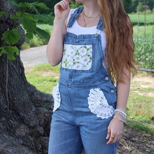 May include: Blue denim overalls featuring a white floral fabric patch on the chest pocket and white crocheted doily accents on the side pockets. The overalls are worn over a plain white tank top, showcasing a medium wash denim finish.