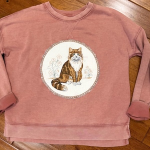 Könnte beinhalten: Ein altrosa Langarm-Sweatshirt mit einer kreisförmigen Grafik auf der Brust. Die Grafik zeigt eine braun-weiße Langhaarkatze, die zwischen einfach gezeichneten Blumen sitzt, eingefasst von einem dekorativen Rand mit Wellenmuster.