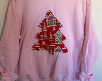 May include: A light pink long-sleeve sweatshirt featuring a large, tree-shaped fabric appliqué centred on the chest. The appliqué is made of red fabric with a repeating pattern of gingerbread houses, gingerbread people, and trees.