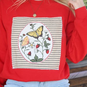 May include: A bright red long-sleeve sweatshirt featuring a large, rectangular fabric patch on the chest. The patch has a beige and black striped background with an oval illustration of two butterflies, white flowers, and red strawberries.
