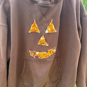 Könnte beinhalten: Ein brauner Kapuzenpullover mit einem Kürbisgesicht-Design auf der Vorderseite. Das Gesicht besteht aus drei orangefarbenen und gelben, strukturierten dreieckigen Aufnähern für Augen und Nase sowie einem gezackten Aufnäher für den Mund.