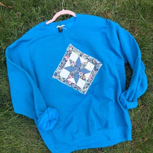 May include: A bright blue long-sleeve sweatshirt featuring a diamond-shaped fabric patch on the chest. The patch displays a star pattern composed of solid blue, white, and floral-printed fabric squares with frayed, raw edges.