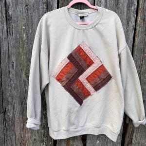 May include: A cream-colored crewneck sweatshirt featuring a large, diamond-shaped patch on the front. The patch is constructed from strips of fabric in shades of brown, rust orange, and light pink, arranged in a geometric, woven-style pattern.
