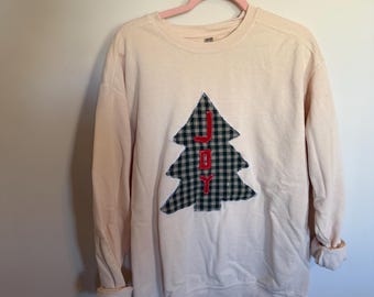 Könnte beinhalten: Ein hellrosa Sweatshirt mit Rundhalsausschnitt und einer zentralen Applikation in Form eines Weihnachtsbaums aus dunkelgrünem und schwarzem Karostoff. Das Wort JOY ist mit rotem Garn vertikal in die Mitte der Applikation gestickt.