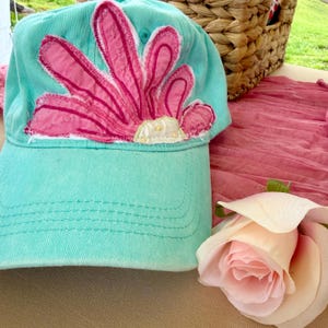 May include: A turquoise baseball cap with a large pink flower applique. The flower has a white and yellow center and is stitched onto the cap. The cap is made of a soft material and has a curved brim.