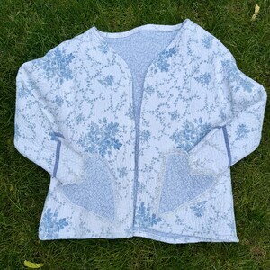 May include: A quilted jacket in white with a blue floral pattern. The jacket has a V-neck and two heart-shaped pockets. The sleeves and edges are trimmed with blue fabric. The jacket is lying on green grass.