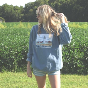 May include: A dusty blue long-sleeve crewneck sweatshirt featuring a rectangular graphic print of a white lighthouse on a rocky coastline with surrounding buildings, paired with light blue and white horizontally striped shorts.