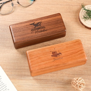 May include: Two rectangular wooden boxes with hinged lids. The darker walnut box features an engraved whale on an open book and the name Nicholas. The lighter cherry-colored box displays an engraved mug on a book and the name Jessica.