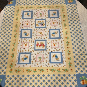 May include: A rectangular fabric featuring a repeating pattern of bunnies in blue and pink squares, set against a white background with small brown shapes. The border has a yellow and blue checkered design with fruit illustrations.