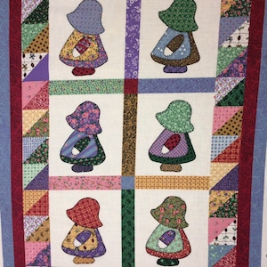 May include: A colorful patchwork quilt featuring four figures in bonnets and aprons. The figures are set against a white background, framed by a border of triangles in various patterns and colors, including pink, green, and yellow. The quilt has a blue border.