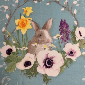 May include: A fabric design featuring a gray and white bunny surrounded by spring flowers, including yellow daffodils, white anemones with purple centers, and purple wisteria. The background is a light teal color with a decorative border.