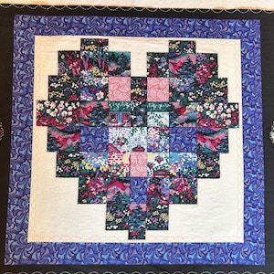 May include: A rectangular fabric quilt featuring a central heart motif composed of small, multi-colored floral and landscape patterned squares. The heart is set against a cream-colored background, framed by a blue patterned border and black fabric.