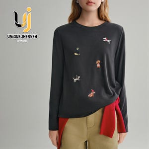 May include: A charcoal grey long-sleeved top adorned with embroidered dog motifs. The top has a round neck and is worn with khaki trousers and a red sweater tied at the waist. The brand name "UNIQUEJHERSEY APPAREL" is visible on the upper left.