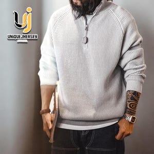 Può includere: Un maglione a coste grigio chiaro con chiusura a zip a un quarto. Il maglione ha un collo alto e maniche lunghe. Il logo del marchio "UNIQUEJHERSEY APPAREL" è visibile in alto a sinistra.