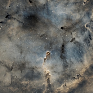 Könnte beinhalten: Ein Himmelsbild, das einen Nebel mit wirbelnden Gas- und Staubwolken zeigt. Die Farben reichen von tiefem Blau und Grau bis hin zu Gold- und Brauntönen. Eine markante säulenartige Struktur erhebt sich aus der Mitte, umgeben von Sternen.