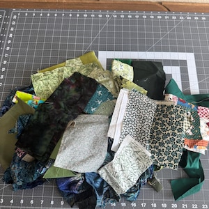 May include: A pile of fabric scraps in various shades of green, blue, and white, with floral and abstract patterns. The fabrics are arranged on a gray cutting mat with inch measurements.