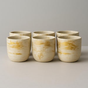 May include: Six cream-colored ceramic cups with a gold metallic swirl design. The cups have a rounded shape and a smooth, matte finish. The gold accents add a touch of elegance to the simple design. Perfect for serving beverages.