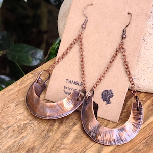 May include: A pair of copper earrings with a hammered finish and a crescent moon shape. The earrings hang from a delicate chain with a hook closure.
