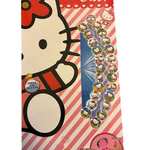 May include: A Hello Kitty-themed toy with a red and white striped background. The packaging features a large Hello Kitty graphic, the text "Hello Cat!", and several small circular images of Hello Kitty. The toy includes a projection switch and the text "Press the Button".