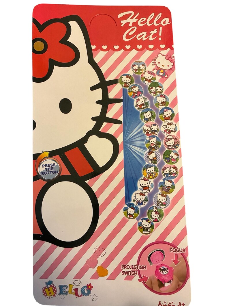 May include: A Hello Kitty-themed toy with a red and white striped background. The packaging features a large Hello Kitty graphic, the text "Hello Cat!", and several small circular images of Hello Kitty. The toy includes a projection switch and the text "Press the Button".